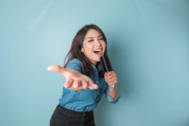 Portrait of carefree Asian woman, having fun karaoke, singing in microphone while standing over blue background