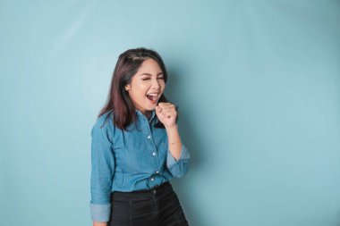 Portrait of carefree Asian woman, having fun karaoke, singing in microphone while standing over blue background