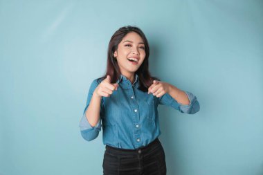 Young beautiful Asian girl wearing a casual blue shirt standing over isolated blue background pointing fingers at the camera with a happy and funny face. Good energy and vibes.