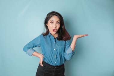 Shocked Asian woman wearing blue shirt pointing at the copy space beside her, isolated by blue background