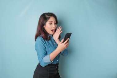 Surprised Asian woman wearing blue shirt pointing at her smartphone, isolated by blue background