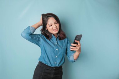 Tired thinking woman, has a sad expression while holding her smartphone. Isolated blue background