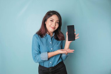 Excited Asian woman wearing blue shirt pointing at the copy space on her smartphone, isolated by blue background