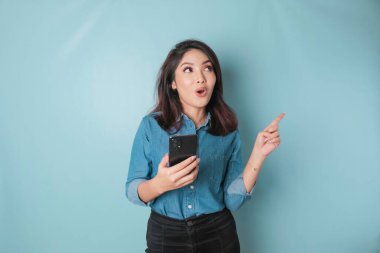 Surprised Asian woman wearing blue shirt pointing at the copy space beside her while holding a smartphone, isolated by blue background