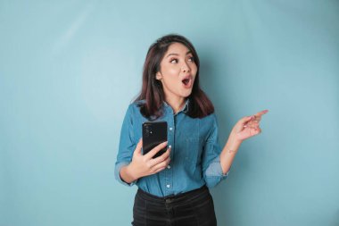 Surprised Asian woman wearing blue shirt pointing at the copy space beside her while holding a smartphone, isolated by blue background