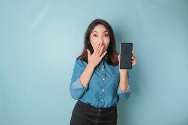 Surprised Asian woman wearing blue shirt pointing at her smartphone, isolated by blue background