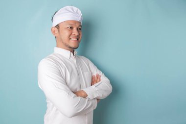 Portrait of a confident smiling Balinese man standing with arms folded and looking at the camera isolated over blue background, wearing a blue shirt
