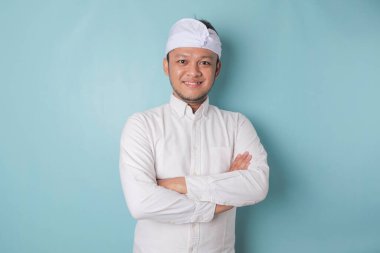 Portrait of a confident smiling Balinese man standing with arms folded and looking at the camera isolated over blue background, wearing a blue shirt