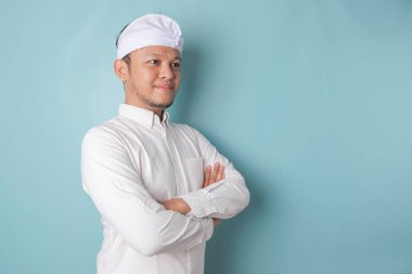 Portrait of a confident smiling Balinese man standing with arms folded and looking at the camera isolated over blue background, wearing a blue shirt