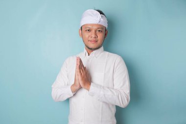 Smiling young Balinese man wearing udeng or traditional headband and white shirt gesturing greeting or namaste isolated over blue background