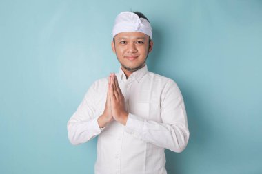 Smiling young Balinese man wearing udeng or traditional headband and white shirt gesturing greeting or namaste isolated over blue background
