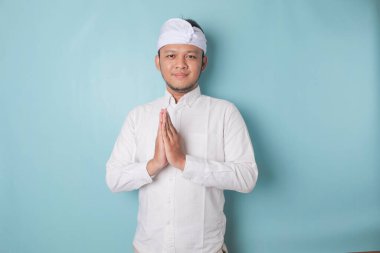 Smiling young Balinese man wearing udeng or traditional headband and white shirt gesturing greeting or namaste isolated over blue background