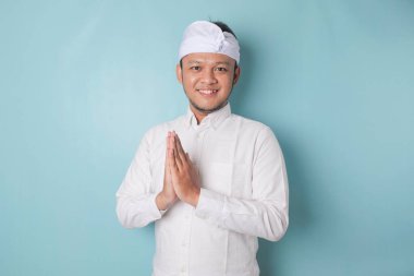 Smiling young Balinese man wearing udeng or traditional headband and white shirt gesturing greeting or namaste isolated over blue background