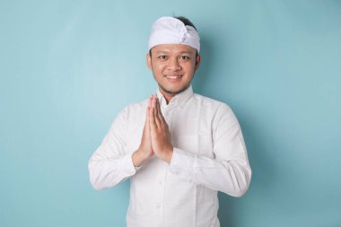 Smiling young Balinese man wearing udeng or traditional headband and white shirt gesturing greeting or namaste isolated over blue background