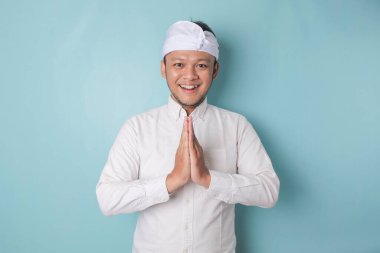 Smiling young Balinese man wearing udeng or traditional headband and white shirt gesturing greeting or namaste isolated over blue background