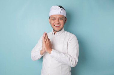 Smiling young Balinese man wearing udeng or traditional headband and white shirt gesturing greeting or namaste isolated over blue background
