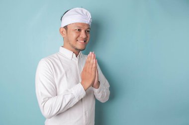 Smiling young Balinese man wearing udeng or traditional headband and white shirt gesturing greeting or namaste isolated over blue background