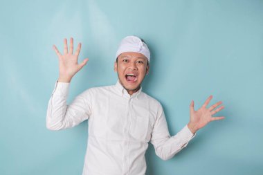 Surprised Balinese man wearing udeng or traditional headband and white shirt, isolated by blue background