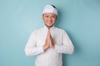 Smiling young Balinese man wearing udeng or traditional headband and white shirt gesturing greeting or namaste isolated over blue background