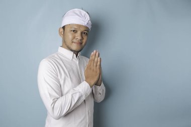 Smiling young Balinese man wearing udeng or traditional headband and white shirt gesturing greeting or namaste isolated over blue background