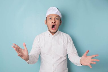 Surprised Balinese man wearing udeng or traditional headband and white shirt, isolated by blue background