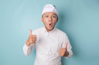 Surprised Balinese man wearing udeng or traditional headband and white shirt, isolated by blue background