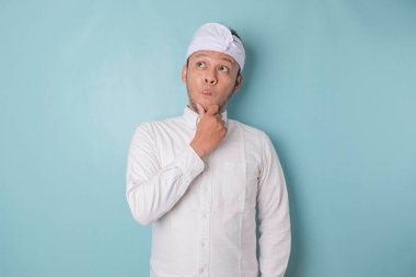 Portrait of a thoughtful young Balinese man wearing udeng or traditional headband and white shirt looking aside while his finger on his chin isolated over blue background