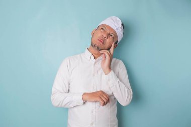 Portrait of a thoughtful young Balinese man wearing udeng or traditional headband and white shirt looking aside while his finger on his chin isolated over blue background
