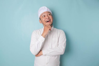 Portrait of a thoughtful young Balinese man wearing udeng or traditional headband and white shirt looking aside while his finger on his chin isolated over blue background