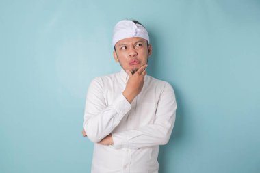 Portrait of a thoughtful young Balinese man wearing udeng or traditional headband and white shirt looking aside while his finger on his chin isolated over blue background