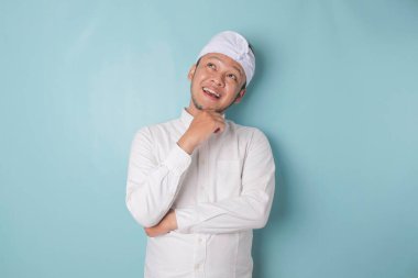 Portrait of a thoughtful young Balinese man wearing udeng or traditional headband and white shirt looking aside while his finger on his chin isolated over blue background