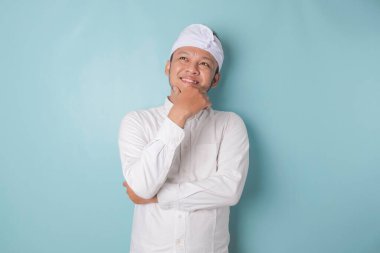 Portrait of a thoughtful young Balinese man wearing udeng or traditional headband and white shirt looking aside while his finger on his chin isolated over blue background