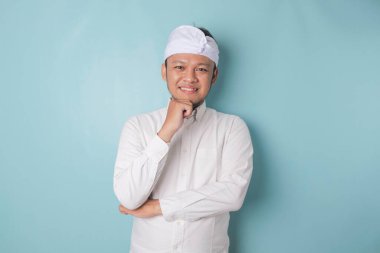 Portrait of a thoughtful young Balinese man wearing udeng or traditional headband and white shirt looking aside while his finger on his chin isolated over blue background
