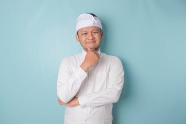 Portrait of a thoughtful young Balinese man wearing udeng or traditional headband and white shirt looking aside while his finger on his chin isolated over blue background