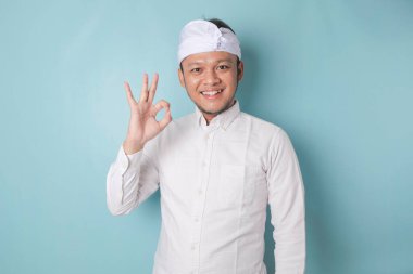 Excited Balinese man wearing udeng or traditional headband and white shirt giving an OK hand gesture isolated by a blue background