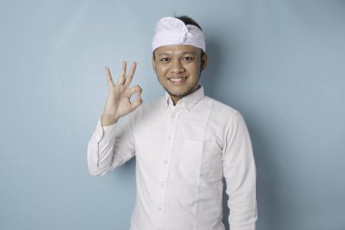 Excited Balinese man wearing udeng or traditional headband and white shirt giving an OK hand gesture isolated by a blue background