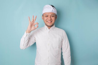 Excited Balinese man wearing udeng or traditional headband and white shirt giving an OK hand gesture isolated by a blue background