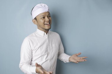 Young male doctor presenting an idea wearing udeng or traditional headband and white shirt while looking smiling on isolated blue background