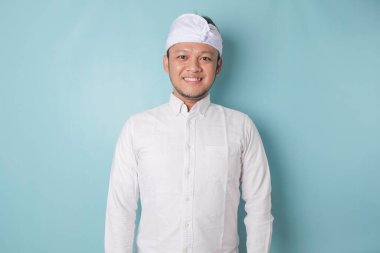 A portrait of a happy Balinese man is smiling wearing udeng or traditional headband and white shirt isolated by a blue background