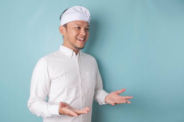 Young male doctor presenting an idea wearing udeng or traditional headband and white shirt while looking smiling on isolated blue background