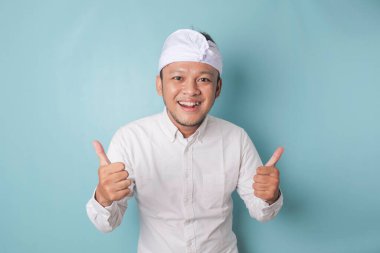 Excited Balinese man wearing udeng or traditional headband and white shirt gives thumbs up hand gesture of approval, isolated by blue background