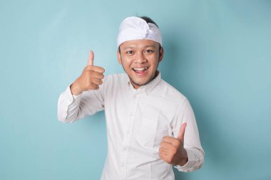 Excited Balinese man wearing udeng or traditional headband and white shirt gives thumbs up hand gesture of approval, isolated by blue background