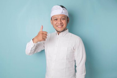 Excited Balinese man wearing udeng or traditional headband and white shirt gives thumbs up hand gesture of approval, isolated by blue background