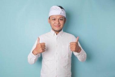 Excited Balinese man wearing udeng or traditional headband and white shirt gives thumbs up hand gesture of approval, isolated by blue background