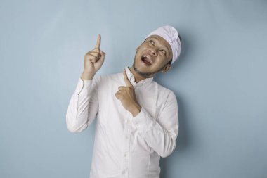 Excited Balinese man wearing udeng or traditional headband and white shirt pointing at the copy space upside him, isolated by blue background