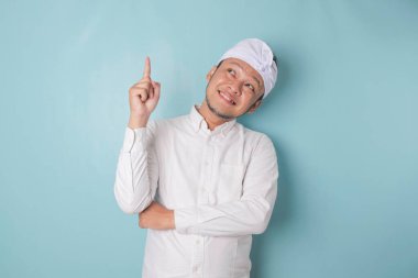 Excited Balinese man wearing udeng or traditional headband and white shirt pointing at the copy space upside him, isolated by blue background