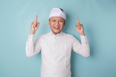 Excited Balinese man wearing udeng or traditional headband and white shirt pointing at the copy space upside him, isolated by blue background
