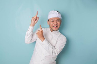 Excited Balinese man wearing udeng or traditional headband and white shirt pointing at the copy space upside him, isolated by blue background