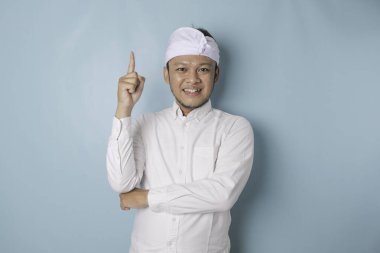 Excited Balinese man wearing udeng or traditional headband and white shirt pointing at the copy space upside him, isolated by blue background