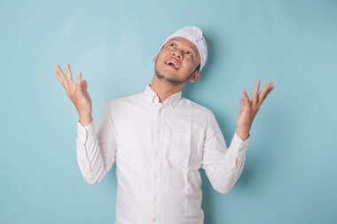 Excited Balinese man wearing udeng or traditional headband and white shirt pointing at the copy space upside him, isolated by blue background
